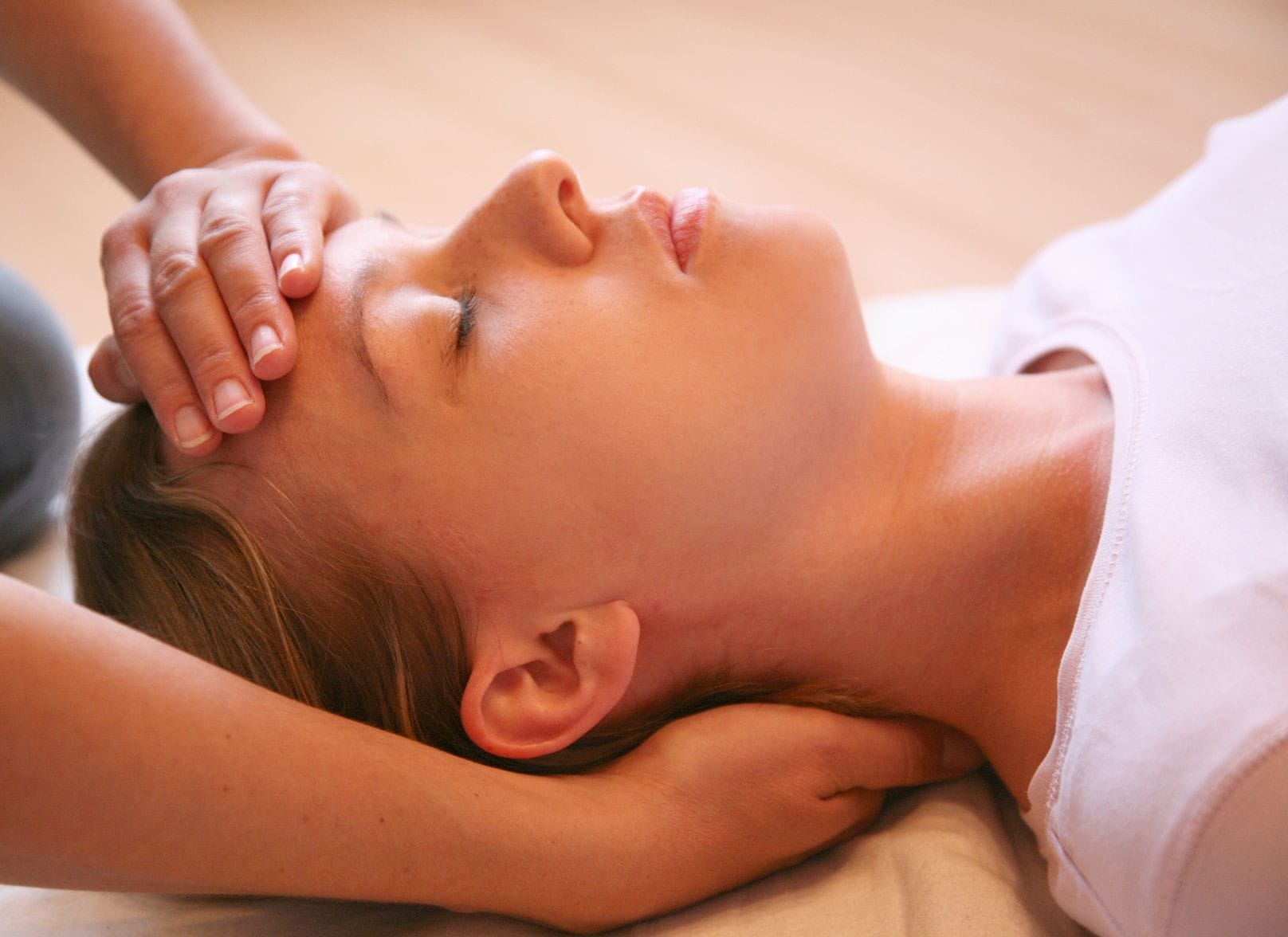 The picture shows a client laying peacefully with the Reiki practitioner placing one hand under the neck and one on the forehead during a session.
