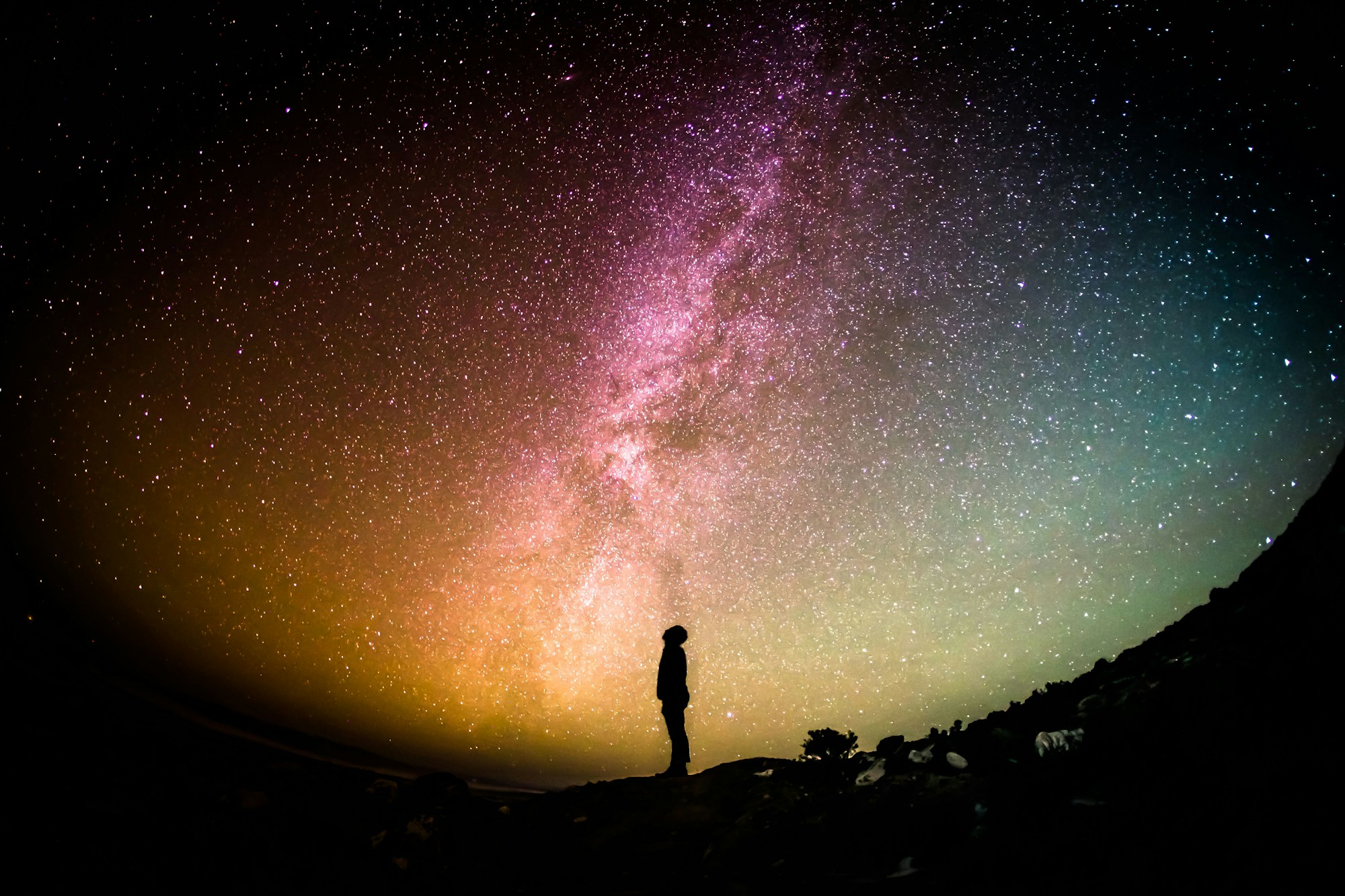 A silhouette of a person is standing in front of a colorful night sky filled with many stars. QHHT helps people connect with the part of us that is the Universe