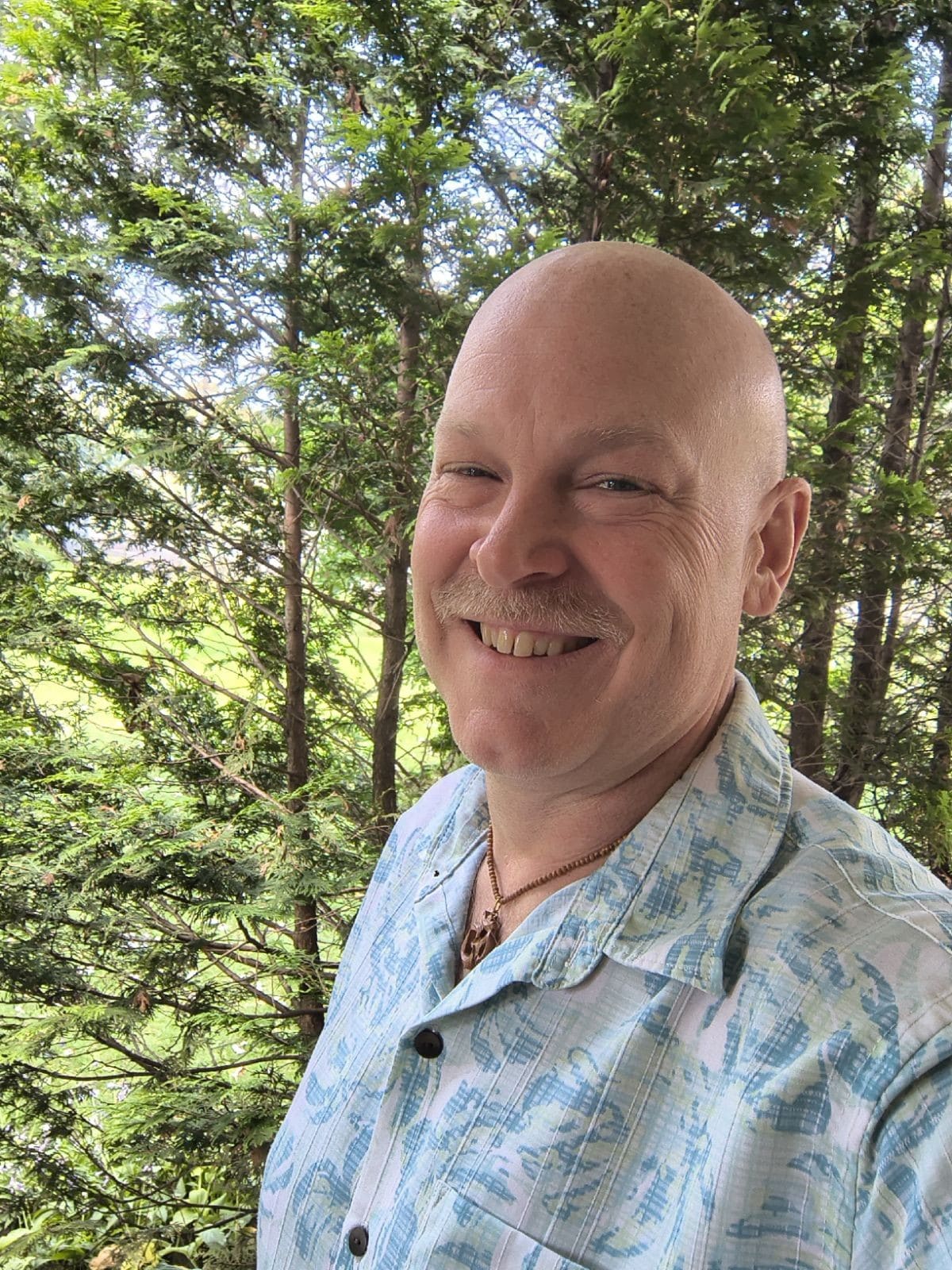 Frank is outside, in front of some pine trees, smiling.
