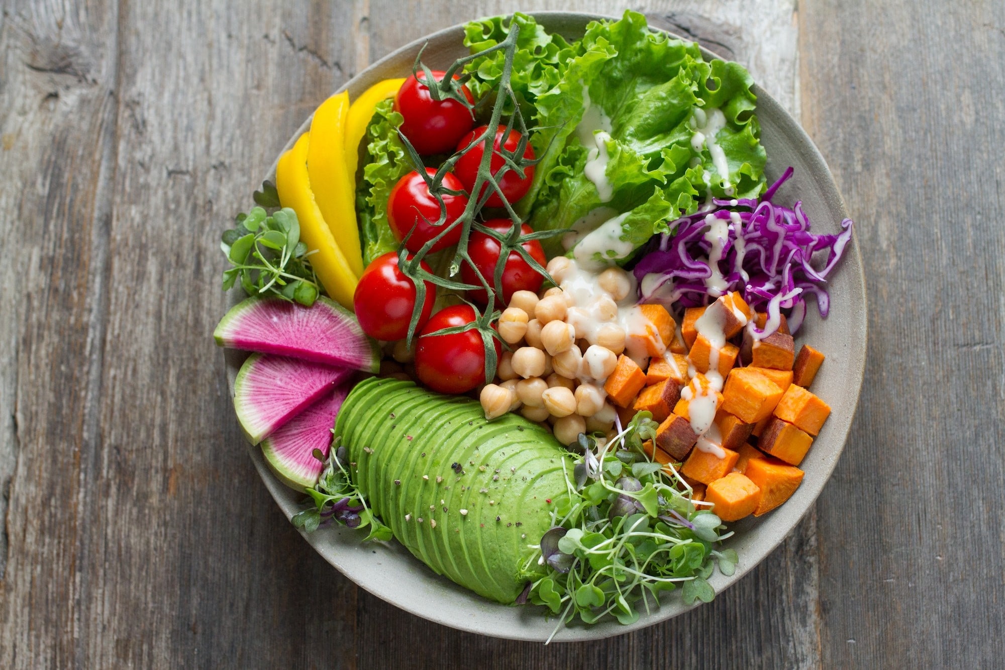 The bowl contains diced squash, purple cabbage, lettuce, yellow pepper slices, cherry tomatoes, avocado, purple vege slices, bean sprouts, and simple dressing.